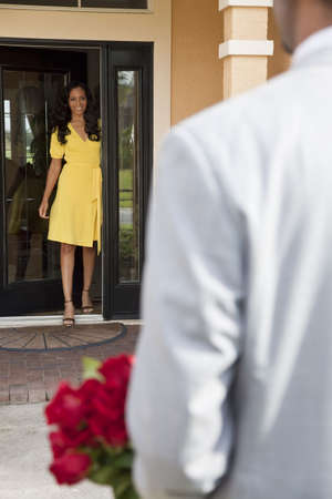A romantic African American man bringing flowers to his happy wife or girlfriend who is standing waiting for him at the door of their home.の写真素材