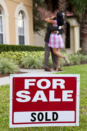 House For Sale and Sold sign with African American couple celebrating the purchase of a house out of focus behind the sign.の写真素材