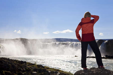 A young woman hiker of explorer shot in golden evening light in front of a huge waterfall. Shot on location at Godafoss waterfall in Iceland, the focus on this shot is on the girl in the foreground.の写真素材