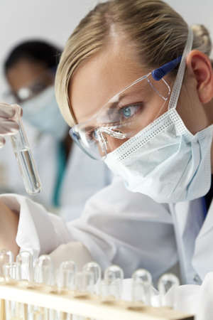A blond medical or scientific researcher or doctor using looking at a clear solution in a laboratory with her Asian female colleague out of focus behind her.の写真素材