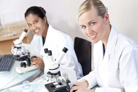 A blond female medical or scientific researcher or doctor using her microscope in a laboratory with her Asian colleague out of focus behind her.の写真素材