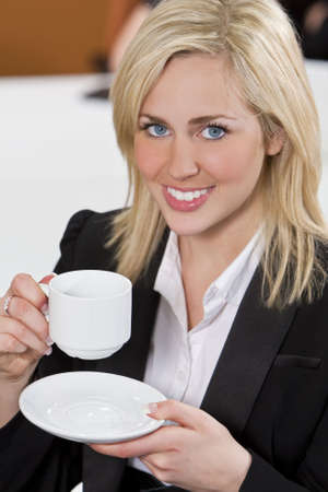 A beautiful young happy blond woman or businesswoman in a suit drinking coffee or tea on a break in an office.の写真素材
