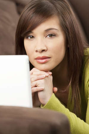 A beautiful young chinese asian oriental woman laying down using a white laptop on her sofa or setteeの写真素材