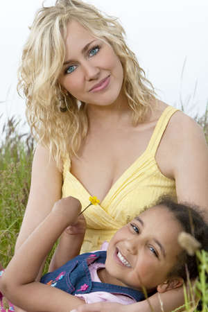A beautiful blond haired blue eyed young woman having fun playing in a field of long grass with a mixed race young girl who is holding a yellow flower.の写真素材