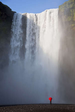 A woman dressed in a red jacket carrying a red umbrella standing at the base of a waterfall. Shot on location at Skogafoss in Iceland.の写真素材