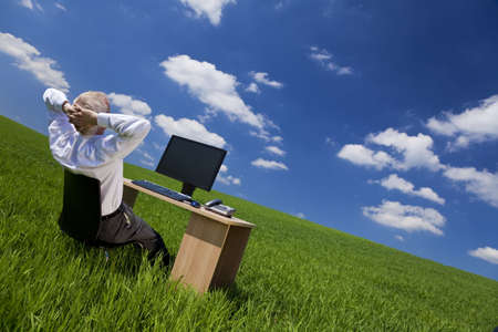 Business concept shot of a middle aged man or businessman relaxing with hands behind his head at an office desk with computer in a green field with a bright blue sky. Shot on location.の写真素材