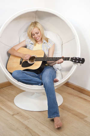 A beautiful young blond woman playing an acoustic guitar in a white bubble chairの写真素材