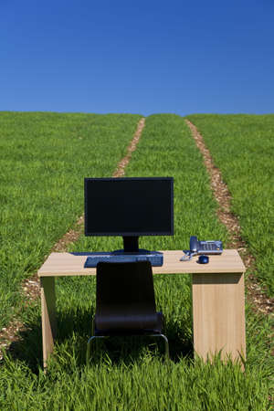 Business concept shot showing a computer on a desk in a green field with a blue sky and a path leading over the horizon. Shot on location not in a studio.の写真素材