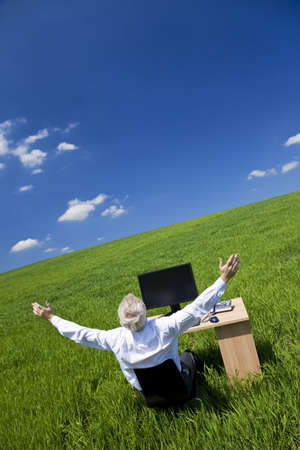 Business concept shot showing an older male executive arms raised using a computer in a green field with a blue sky complete with fluffy white clouds. Shot on location not in a studio.の写真素材
