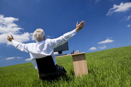 Business concept shot showing an older male executive arms raised using a computer in a green field with a blue sky complete with fluffy white clouds. Shot on location not in a studio.の写真素材
