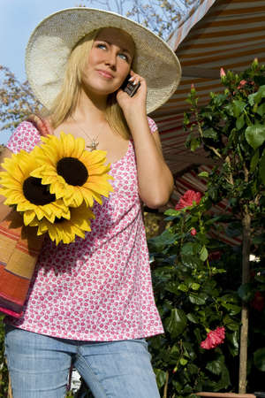 A beautiful young blond woman wearing a sun hat talking on her cell phone while carrying a shopping bag of sunflowers の写真素材