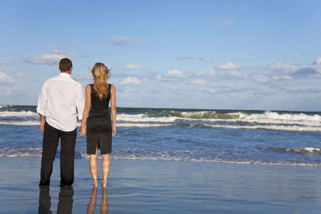 A young man and woman holding hands as a romantic couple looking out to sea on a beach with a bright blue skyの写真素材
