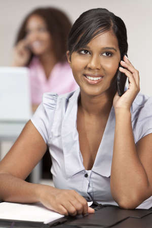 A beautiful young Indian Asian business woman with a wonderful smile in an office talking on her cell phone with her female colleague out of focus behind her.の写真素材