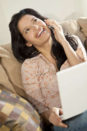 A stunningly beautiful young Latin Hispanic woman sitting on a settee having fun on her laptop and laughing while talking on her cell phoneの写真素材