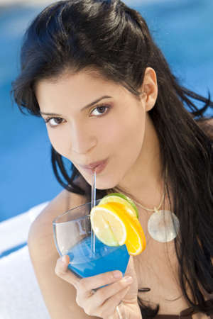 Stunningly beautiful young latina Hispanic woman in drinking a cocktail by a blue swimming poolの写真素材