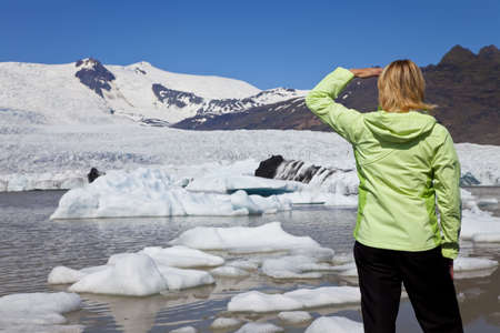 Environmental picture of woman hiker dressed in green jacket looking at the effects of global warming climate change on a melting glacier with icebergs floatiing into a glacial pool.の写真素材