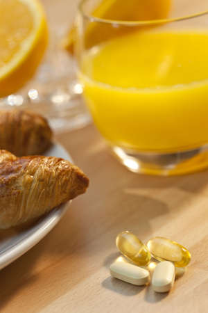 A healthy continental breakfast of croissant pastries, orange juice, oranges and in focus in the foreground are vitamin tablets and cod liver oil capsules, Illuminated with golden early morning sunshine.の写真素材
