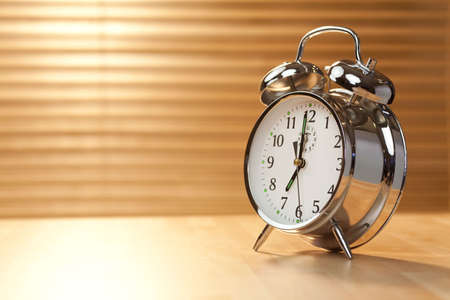 A classic alarm clock illumintaed with golden early morning sunshine coming in through the blind on a window in the background.の写真素材