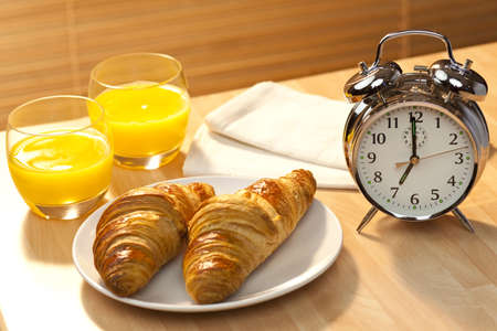 A healthy continental breakfast of croissant pastries, orange juice, oranges Illuminated with golden early morning sunshine and accompanied by a classic alarm clock set at 7amの写真素材