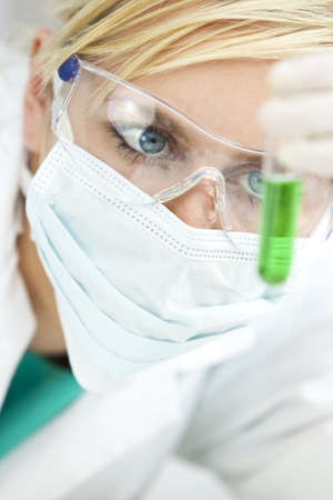 A blond medical or scientific researcher or doctor looking at a test tube of green liquid in a laboratory while wearing surgical mask and safety glasses.の写真素材