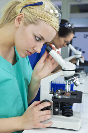 A blond female medical or scientific researcher or doctor using her microscope in a laboratory with her  colleague out of focus behind her.の写真素材