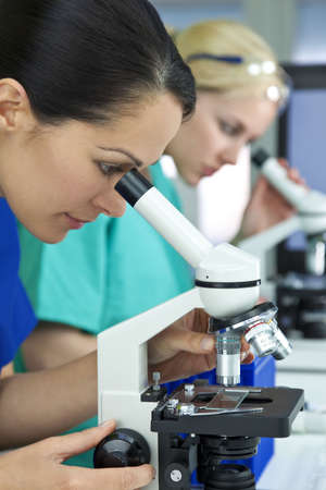 A female medical or scientific researcher or doctor using her microscope in a laboratory with her blond colleague out of focus behind her.の写真素材