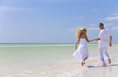 Happy young man and woman couple running, laughing and holding hands on a deserted tropical beach with bright clear blue skyの写真素材