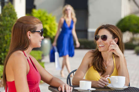 Two beautiful and sophisticated young women friends wearing sunglasses and having coffee around a modern city cafe table With their friend arriving with shopping bags in the backgroundの写真素材