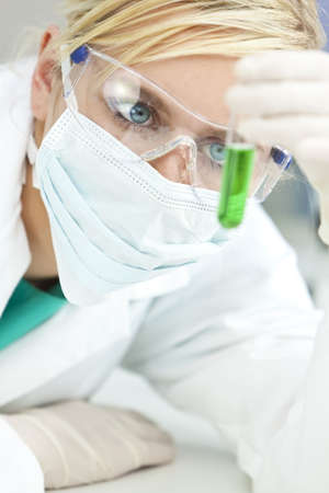 A blond medical or scientist researcher or doctor looking at a test tube of green solution in a laboratory while wearing safety glasses, surgical face mask and gloves.の素材 [FY3107263330]