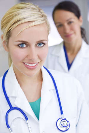 An attractive smiling female doctor wearing a white coat and stethoscope in a hospital with her colleague out of focus behind her.の写真素材