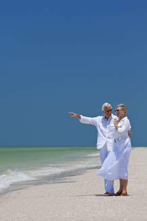 Happy senior man and woman couple together looking out to sea on a deserted tropical beach with bright clear blue sky, the man is pointing to the horizonの写真素材