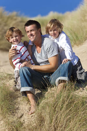 A man and two young boys, father and sons, sitting down and having fun in the sand dunes of a sunny beachの写真素材