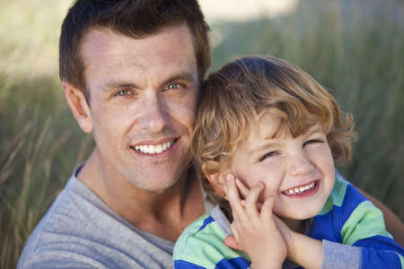 A man and young boy, father and son, sitting down and having fun in the grassy sand dunes of a sunny beachの写真素材