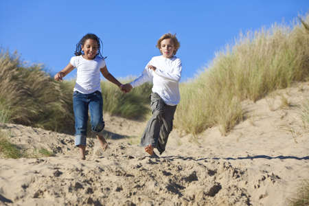 A young mixed race African American girl and a little blond boy running, holding hands and having fun in the sand dunes of a sunny beachの写真素材