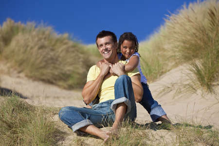 A man and young girl, father and mixed race daughter, playing and having fun in the sand dunes of a sunny beachの写真素材