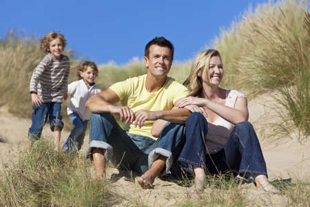 A happy family of mother, father and two sons, sitting and having fun in the sand dunes of a sunny beachの写真素材