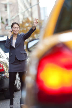 A young woman or businesswoman hailing a yellow Taxi cab while talking on her cell phone in a modern cityの写真素材