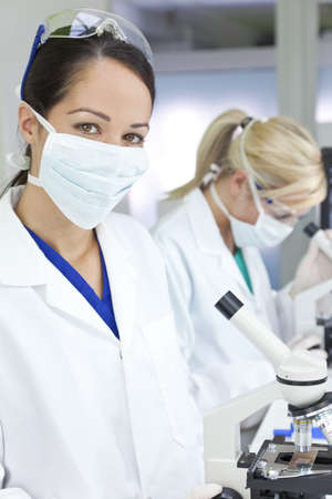 A female medical or scientific researcher or doctor using her microscope in a laboratory with her  colleague out of focus behind her.の写真素材