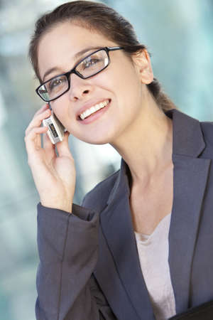 A beautiful smart young woman or businesswoman wearing glasses talking on cell phone in a sunny city location の写真素材