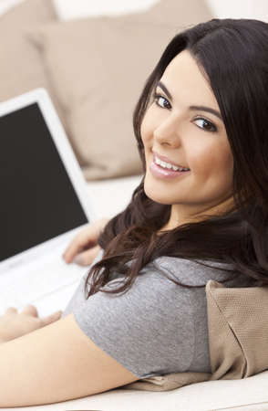 Beautiful happy young Latina Hispanic woman smiling and using a laptop computer at home on her sofaの写真素材