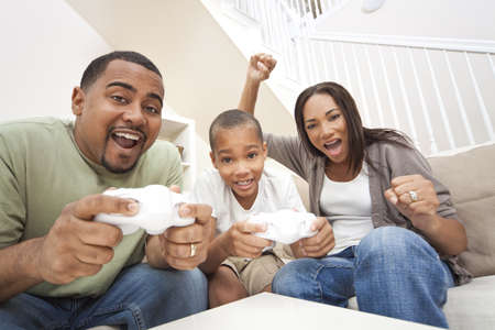 African American family, parents and son, having fun playing computer console games together, Father and son have the handset controllers and the mother is cheering the players.の写真素材