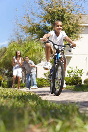 A young African American family with boy child riding his bicycle and his happy excited parents giving encouragement behind himの写真素材