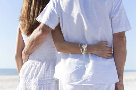 Rear view of man and woman romantic couple in white clothes on a deserted tropical beach with bright clear blue skyの写真素材