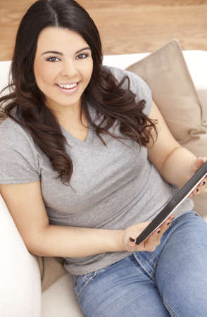 Beautiful happy young Latina Hispanic woman smiling and using a tablet computer at home on her sofaの写真素材