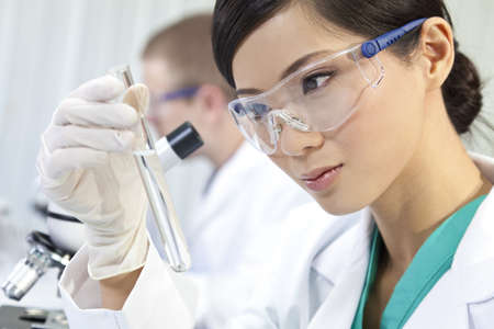 A Chinese Asian female medical or scientific researcher or doctor using looking at a test tube of clear liquid in a laboratory with her colleague out of focus behind her. A Chinese Asian female medical or scientific researcher or doctor using looking at a test tube of clear liquid in a laboratory with her colleague out of focus behind her.