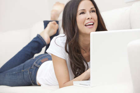 Beautiful happy young Latina Hispanic woman laying down, smiling and using a laptop computer at home on her sofaの写真素材