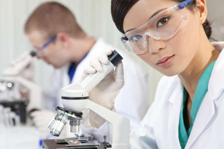 A Chinese Asian female medical or scientific researcher or doctor using a microscope in a laboratory with her colleague out of focus behind her.の写真素材