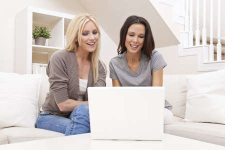 Two beautiful young women at home sitting on sofa or settee using a laptop computer and smilingの写真素材