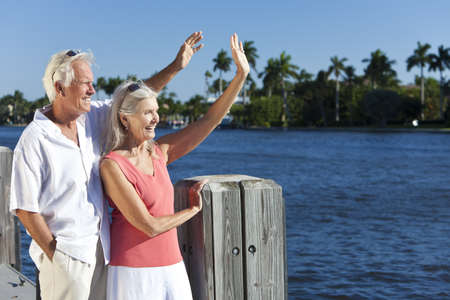 Happy senior man and woman couple together outside in sunshine waving by the sea on a jetty or pierの写真素材