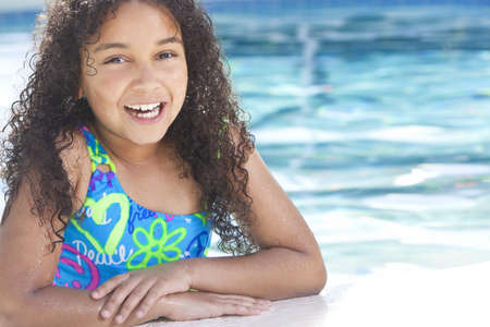 A cute happy young interracial African American girl child relaxing on the side of a swimming pool smiling の写真素材